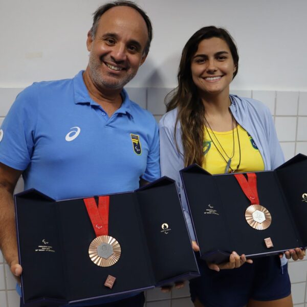 Técnico Leonardo Miglinas e a paratleta Mariana Gesteira exibem as duas medalhas de bronze conquistadas por ela na Paralimpíada de Paris 2024, durante um evento no UniSales. O momento celebra a trajetória do treinador e a conquista da atleta, que é três vezes medalhista paralímpica. Ambos estão sorrindo, com as medalhas em destaque, simbolizando o triunfo e a superação no esporte paralímpico.