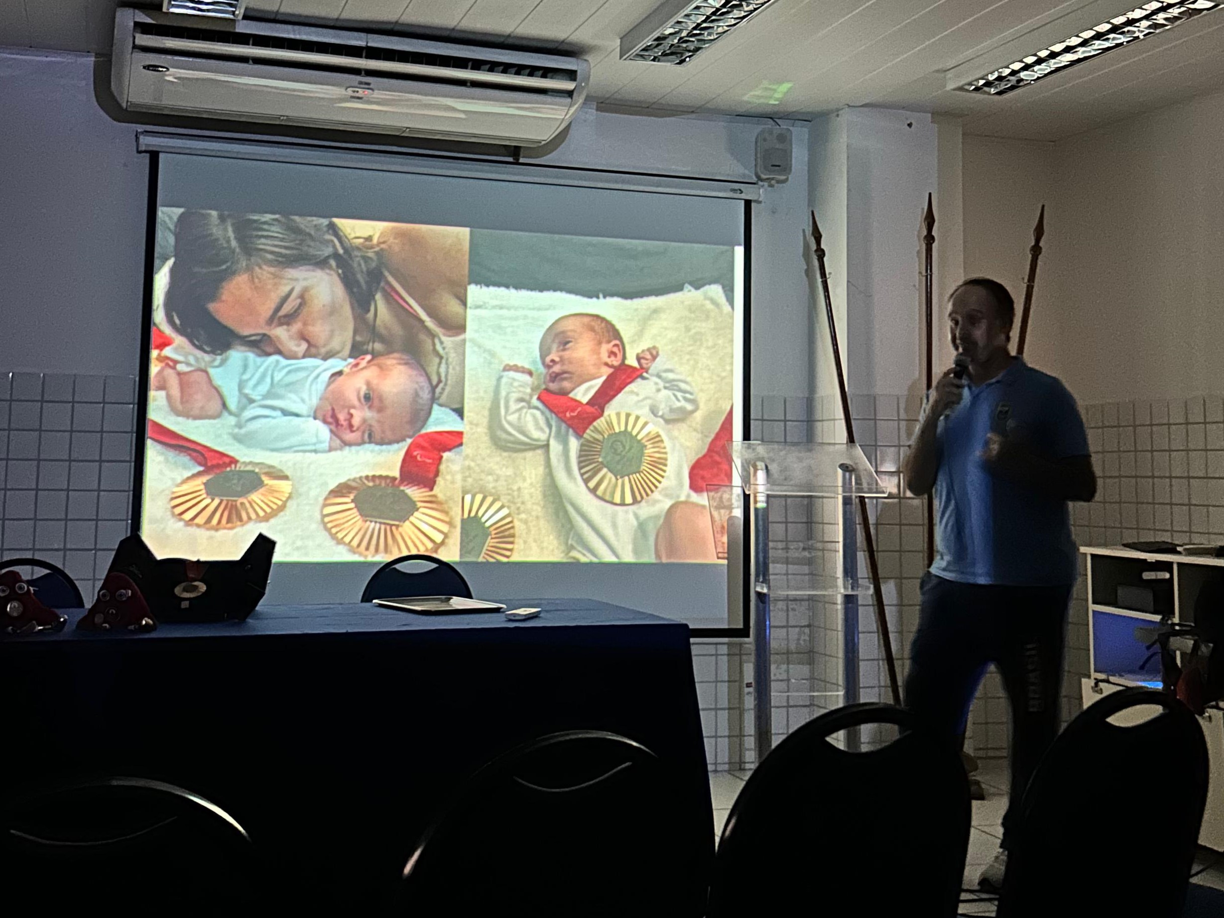  Professor Leonardo Miglinas apresenta um slide que mostra sua filha, Maitê, ao lado da paratleta Mariana Gesteira e uma das medalhas de bronze conquistadas por ela na Paralimpíada de Paris 2024. A imagem simboliza a conexão entre a vida pessoal e as conquistas esportivas, destacando a importância do apoio familiar e as vitórias no esporte paralímpico.