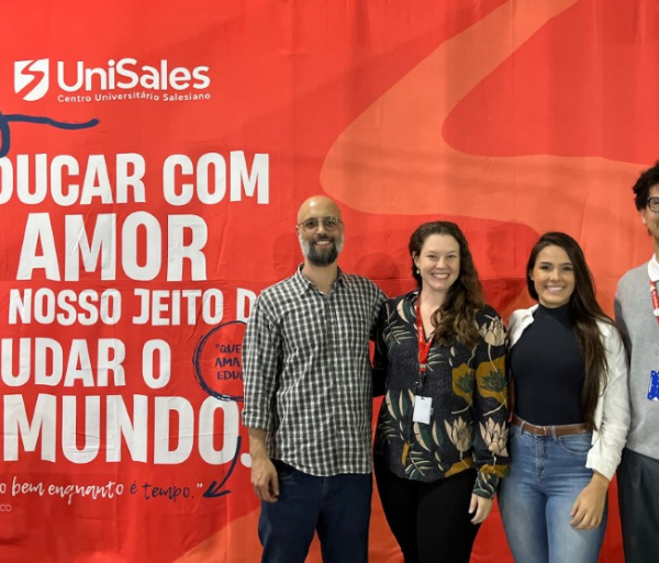 Professores participantes da Semana de Formação de Docentes e Tutores do UniSales 2025/2 posando em frente a painel vermelho com a frase ‘Educar com amor é o nosso jeito de mudar o mundo